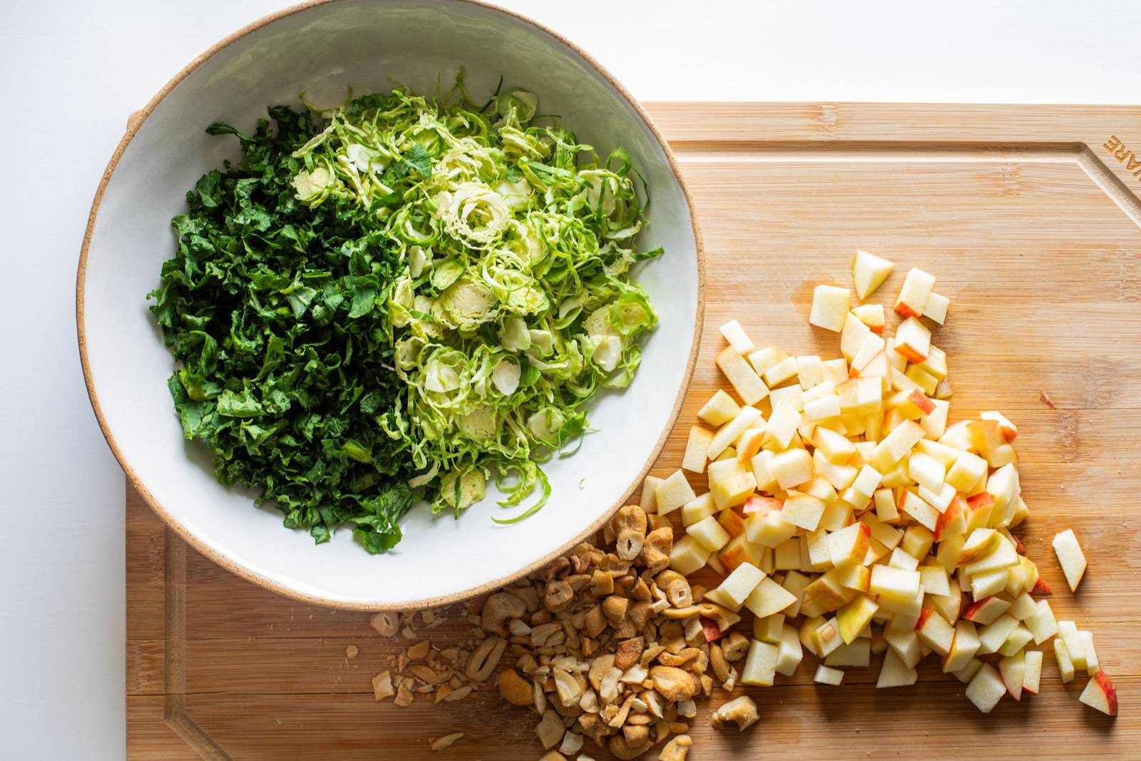 Kale and Brussels Sprouts shredded in a bowl, with apples and cashews chopped on a cutting board.
